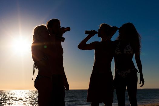 People (two couples) on the beach having a party, drinking and having a lot of fun in the sunset (only silhouette of people to be seen, people having bottles in their hands with the sun shining through)