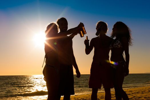 People (two couples) on the beach having a party, drinking and having a lot of fun in the sunset (only silhouette of people to be seen, people having bottles in their hands with the sun shining through)