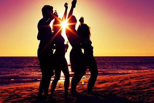 People (two couples) on the beach having a party, drinking and having a lot of fun in the sunset (only silhouette of people to be seen, people having bottles in their hands with the sun shining through)