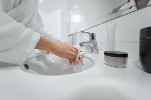 Side view of woman in white robe washing hands with soap in bathroom at home or hotel. Concept of care hands and body for improvements skin with special cosmetics. 