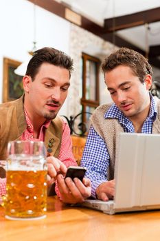 Two friends in Bavarian pub in traditional clothes sitting on a table in a pub with laptop