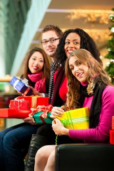 Diversity group of four people - Caucasian, black and Asian - sitting with Christmas presents and bags in a shopping mall in front of a Christmas tree with baubles