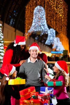 Santa Claus in male and female variants giving Christmas gifts in a shopping mall amid artificial snow covered fir trees and lights