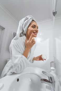 Side view of smiling beautiful young woman looking at mirror and using face cream in white robe. Concept of time for yourself and care body with good mood at home.