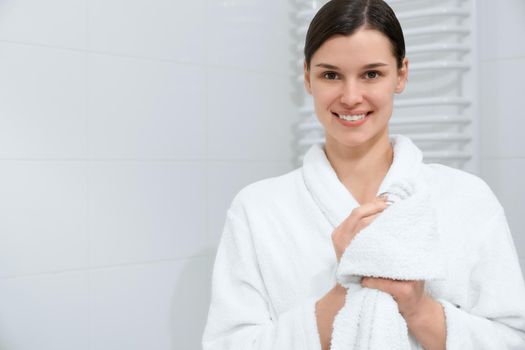 Front view portrait of smiling attractive young woman in white robe holding towel and wiping hands after shower. Concept of time for care body and hands.