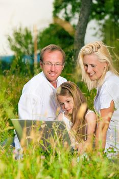 Happy family with daughter girl sitting in a meadow in summer with laptop