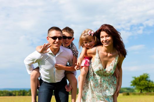 Happy family - mother, father, children - standing on a meadow in summer piggyback the kids