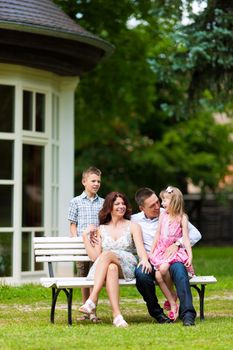 Young happy family sitting in the sun in front of their new home on a bench - it is a villa