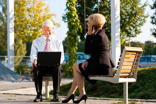 Business people working outdoors - he is working with laptop, she is calling someone on phone