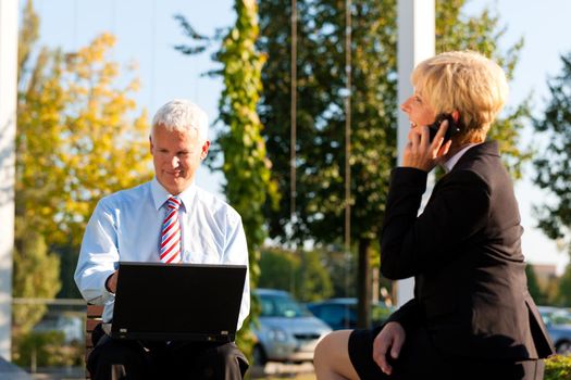 Business people working outdoors - he is working with laptop, she is calling someone on phone