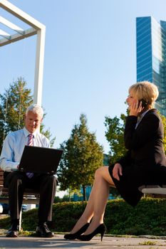 Business people working outdoors - he is working with laptop, she is calling someone on phone