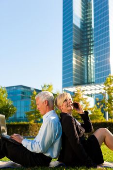 Business people working outdoors - he is working with laptop, she is calling someone on phone