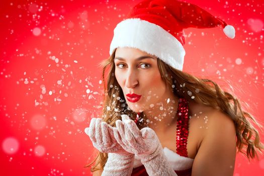 Attractive young woman in Santa Claus costume blowing snow flakes on isolated red background Xmas celebration