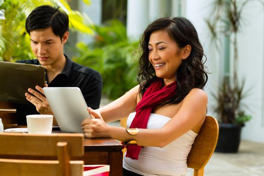 Asian woman and man are sitting in a bar or cafe outdoor and are surfing the internet with a tablet computer