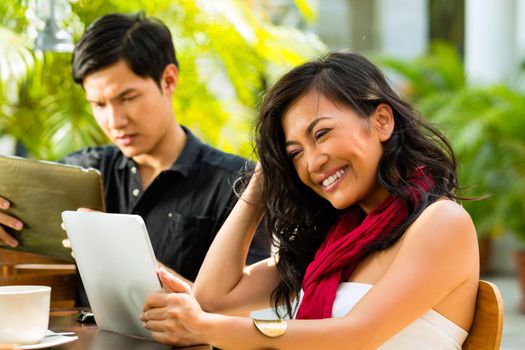 Asian woman and man are sitting in a bar or cafe outdoor and are surfing the internet with a tablet computer