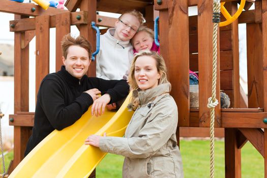 Young Family on the playground, the children sitting on a climbing frame, mother and father standing in front