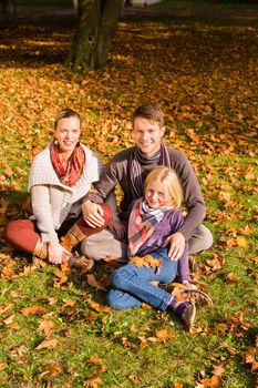 Happy family, Mother, father and daughter or child sitting outdoor on meadow with colorful leaves and under the trees in autumn or fall