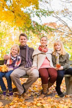 Happy family, Mother, father and daughters or children sitting outdoor on bench with colorful leaves and under the trees in autumn or fall