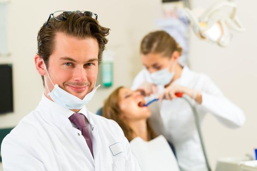 Dentists in his surgery looking at the viewer, in the background his assistant is giving a female patient a treatment