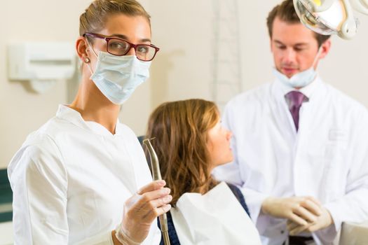 Dentists in his surgery holds a drill and looking at the viewer, in the background her colleague is giving a female patient a treatment