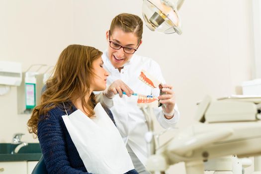 Dentist in his surgery holds a denture and explains a female patient with a toothbrush