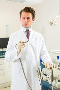 Dentists in his surgery holds a drill and looking at the viewer, in the background are tools for a dentist