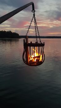 A bonfire burns in a metal basket hanging over the water at sunset