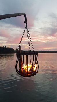 A bonfire burns in a metal basket hanging over the water at sunset