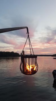 A bonfire burns in a metal basket hanging over the water at sunset