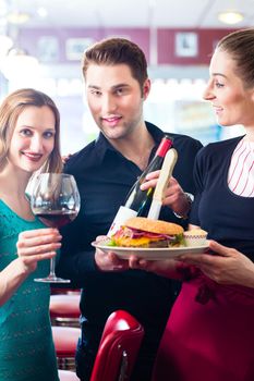 Friends or couple eating fast food in American fast food diner, the waitress serving the food, burgers, fries, and red wine