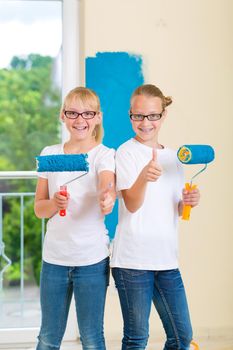 In Family home girl with her sister or friend painting with paint roller a wall in blue