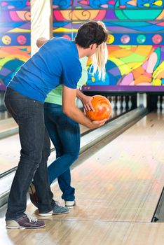 Young couple or friends, man and woman, playing bowling with a ball in front of the ten pin alley, he shows her how it works