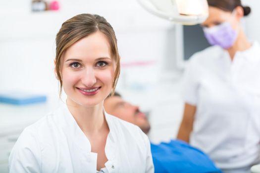 Female Dentist in her surgery looking at the viewer, in the background her assistant is giving a male patient a treatment