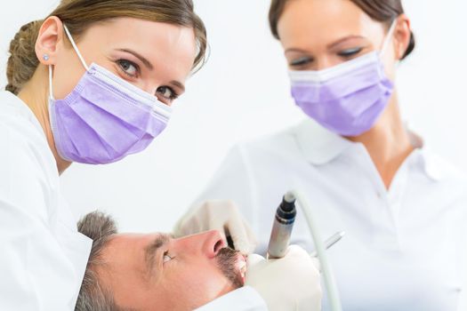 dentist in her practice or office treating male patient with assistant wearing masks and gloves