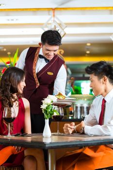 Asian Chinese couple - Man and woman - or lovers having a date or romantic dinner in a fancy restaurant while the waiter is serving food