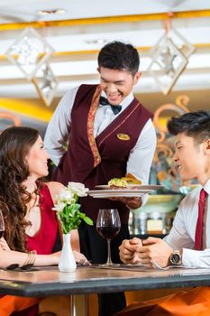Asian Chinese couple - Man and woman - or lovers having a date or romantic dinner in a fancy restaurant while the waiter is serving food