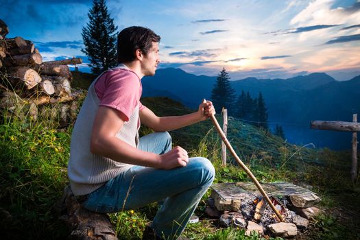Tyrol - Young man sitting on alpine meadow of a mountain on Campfire in the Bavarian Alps and enjoys the romantic evening sunset of the panorama in leisure time or vacation