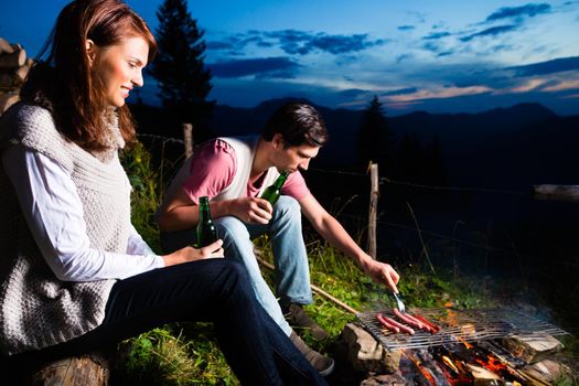 Tyrol - Young couple sitting on alpine meadow of a mountain on Campfire in the Bavarian Alps enjoying the romantic evening sunset of the panorama in leisure time or vacation