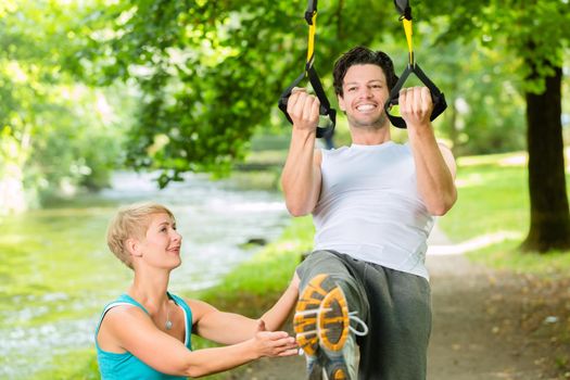 Young fitness man exercising with suspension trainer sling and personal sport trainer in City Park under summer trees