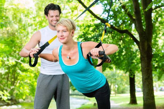Fitness woman exercising with suspension trainer and personal sport trainer in City Park under summer trees