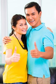 Young Asian handsome couple in apartment with city view showing thumbs up