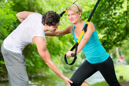 Fitness woman exercising with suspension trainer and personal sport trainer in City Park under summer trees