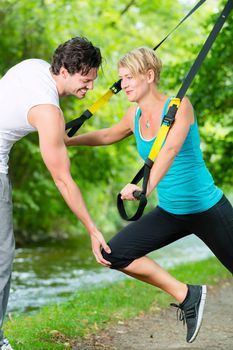 Fitness woman exercising with suspension trainer and personal sport trainer in City Park under summer trees