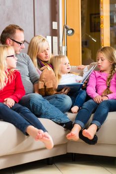 Family sitting together with mother, father and children comfortable on sofa, listening to fairy tale from story book