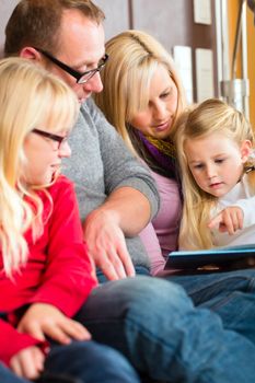 Family sitting together with mother, father and children comfortable on sofa, listening to fairy tale from story book