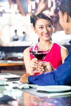 Asian couple having dinner and drinking red wine in very fancy restaurant with open kitchen in background