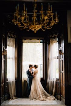 Groom almost kisses bride against the background of the large windows of the old villa. View from the next room with a chandelier with candlesticks on the ceiling. Lake Como. High quality photo