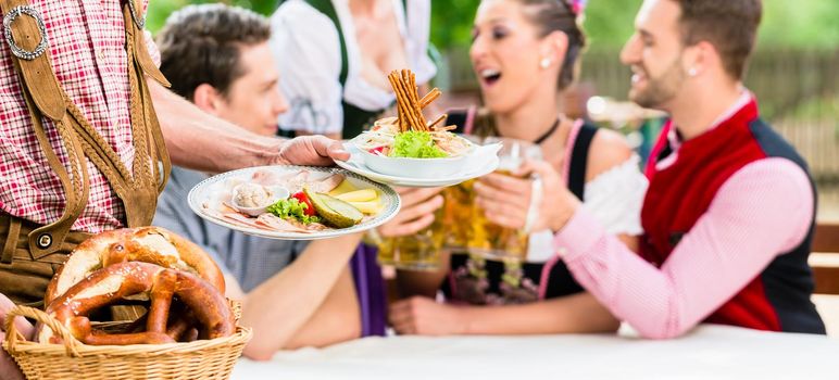 Waiter serving food in Bavarian beer garden, people eating and drinking in background