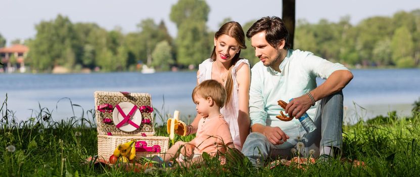 Family having picnic at lake sitting on meadow eating and drinking
