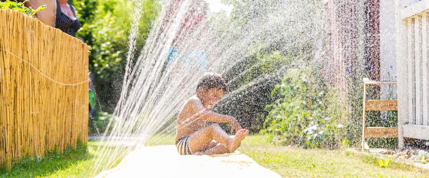 Boy cooling down with garden hose, family in the background on a hot summer day